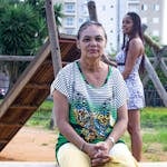 Elderly woman sitting in a park in Brazil, with greenery and playground background.